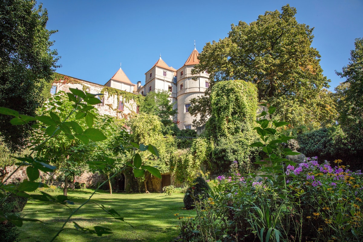 A scenic outdoor view showcases a well-maintained garden featuring diverse greenery and flowering plants. In the background, a historic building with distinctive towers stands amidst trees, offering a sense of tranquility and history.