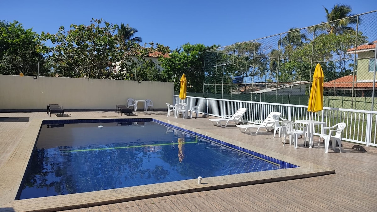 A refreshing swimming pool is framed by wooden decking and surrounded by lounge chairs. Sun umbrellas provide shade, while palm trees and greenery create a serene backdrop. The area is well-lit under clear blue skies, suggesting a perfect space for relaxation.