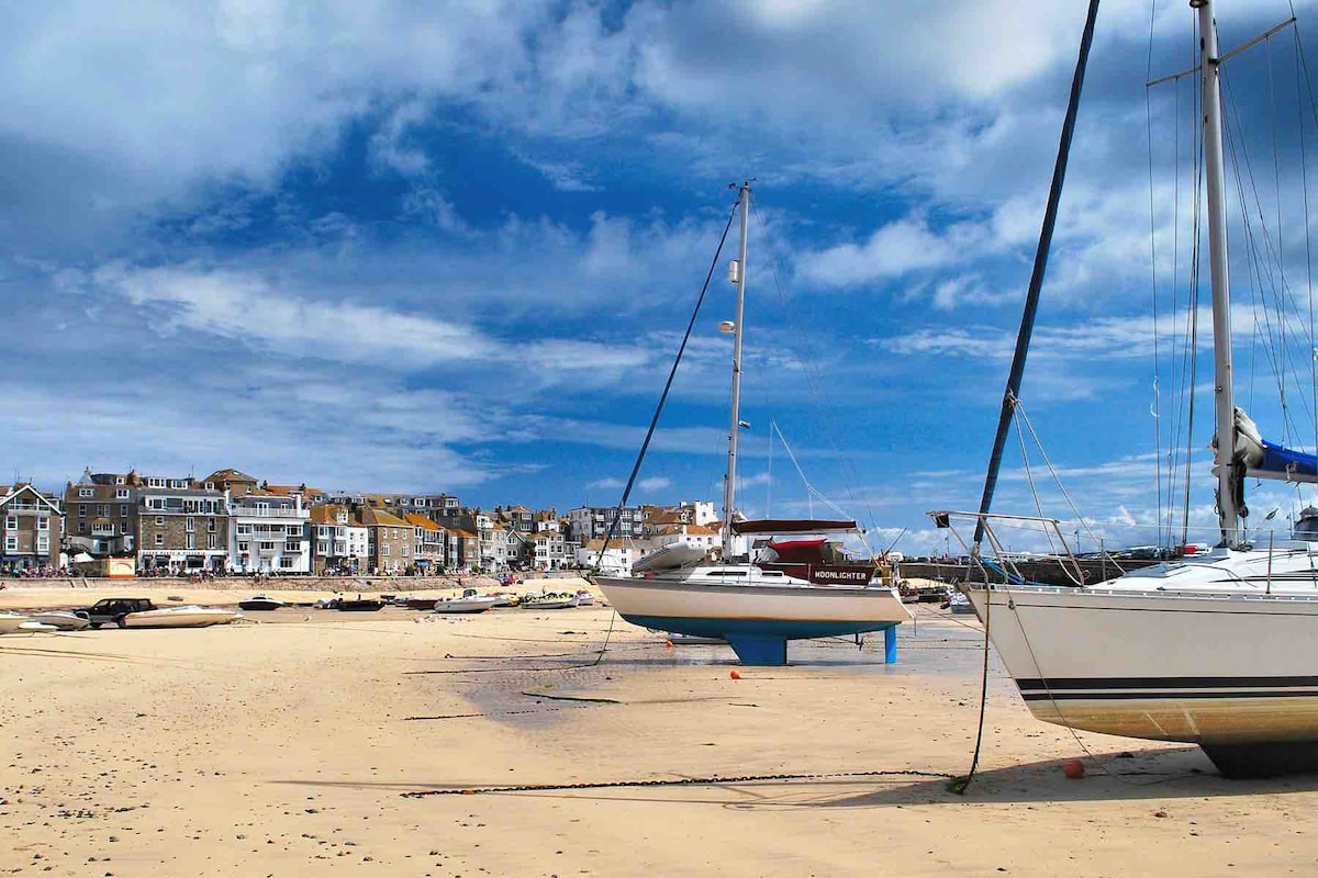 A sandy beach is pictured with sailboats anchored in shallow water. The sky is partially cloudy, casting gentle shadows. In the background, buildings line the shore, showcasing a mix of architectural styles. The scene conveys a coastal atmosphere, ideal for relaxation and exploration.