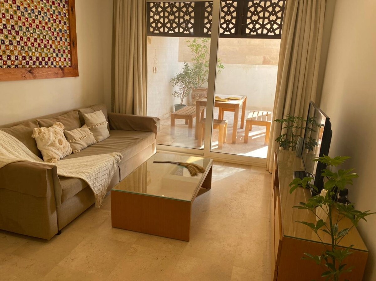 A living room features a spacious beige sofa and a glass coffee table, complemented by wooden accents. Natural light enters through large sliding doors, leading to a terrace area with outdoor seating. A television is positioned on a wooden console, enhancing the cozy setting.