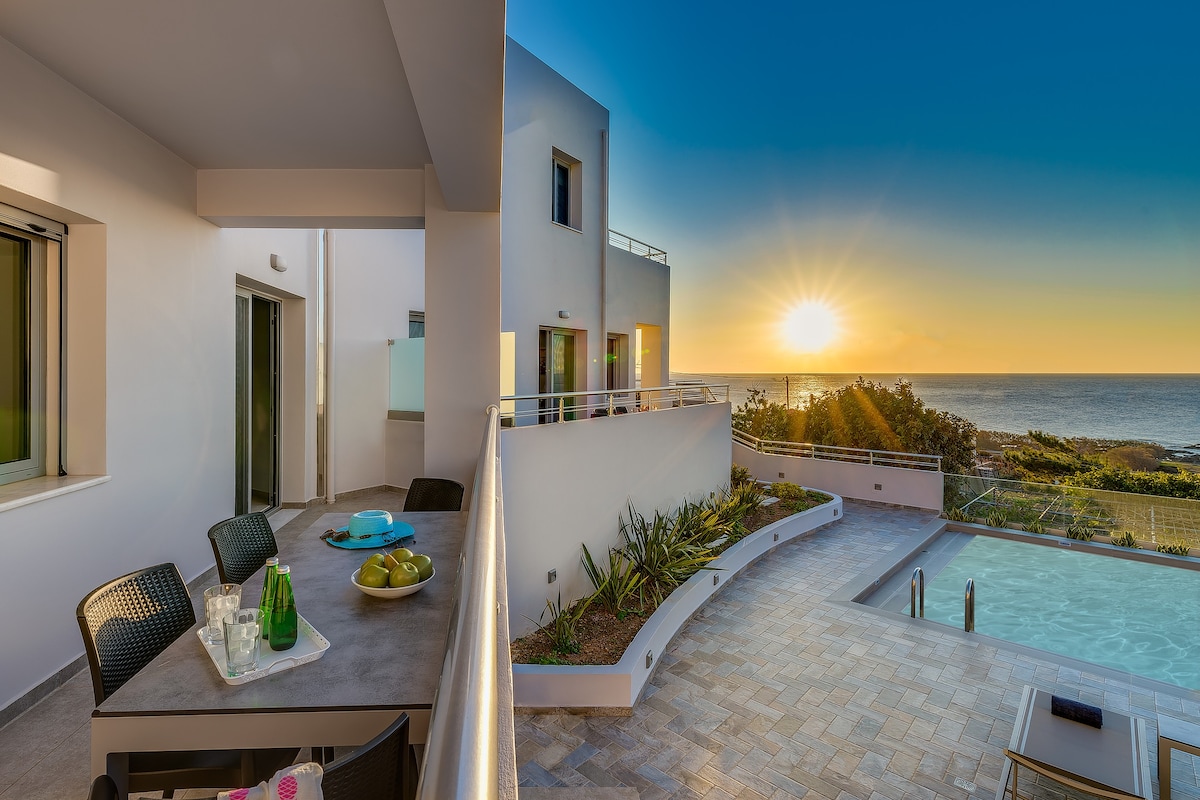 A balcony area showcases a dining table set with a green water pitcher and bowls of fruit. Beyond, a tranquil pool area is visible, with sun setting behind the serene sea, illuminating the sky in warm tones.
