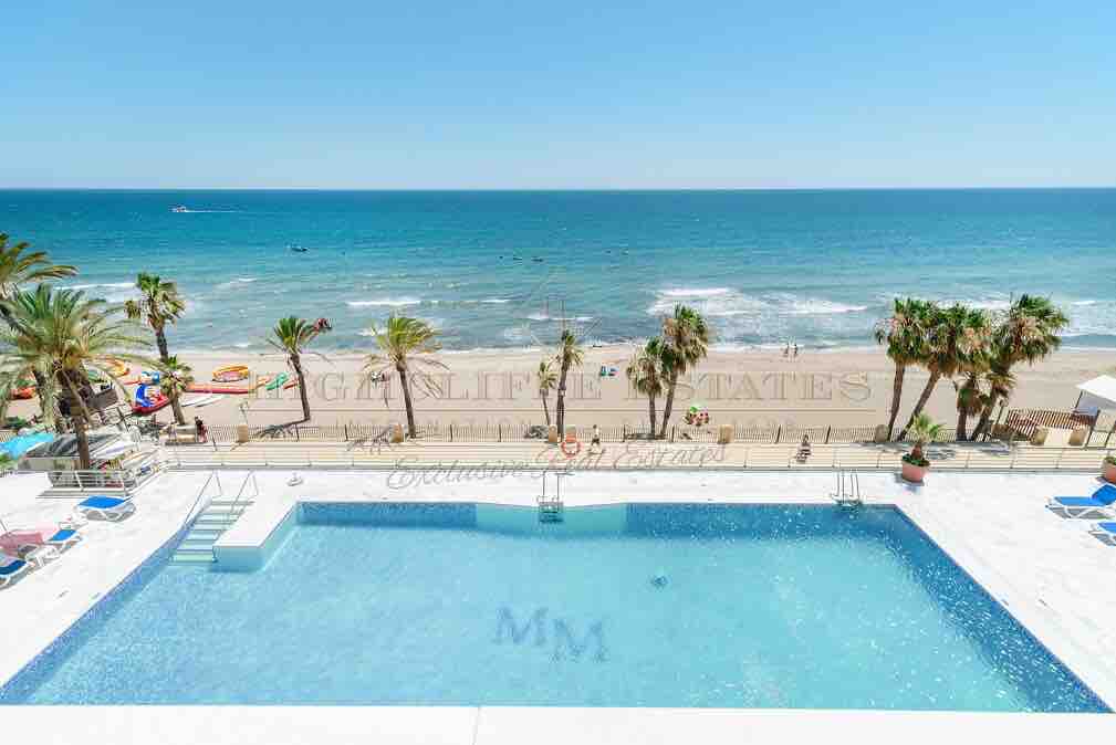 An outdoor pool is visible in the foreground, reflecting the clear blue sky. Beyond the pool, the beach stretches with waves gently lapping at the shore. Palm trees sway beside colorful sun loungers, creating a relaxed atmosphere by the seaside.