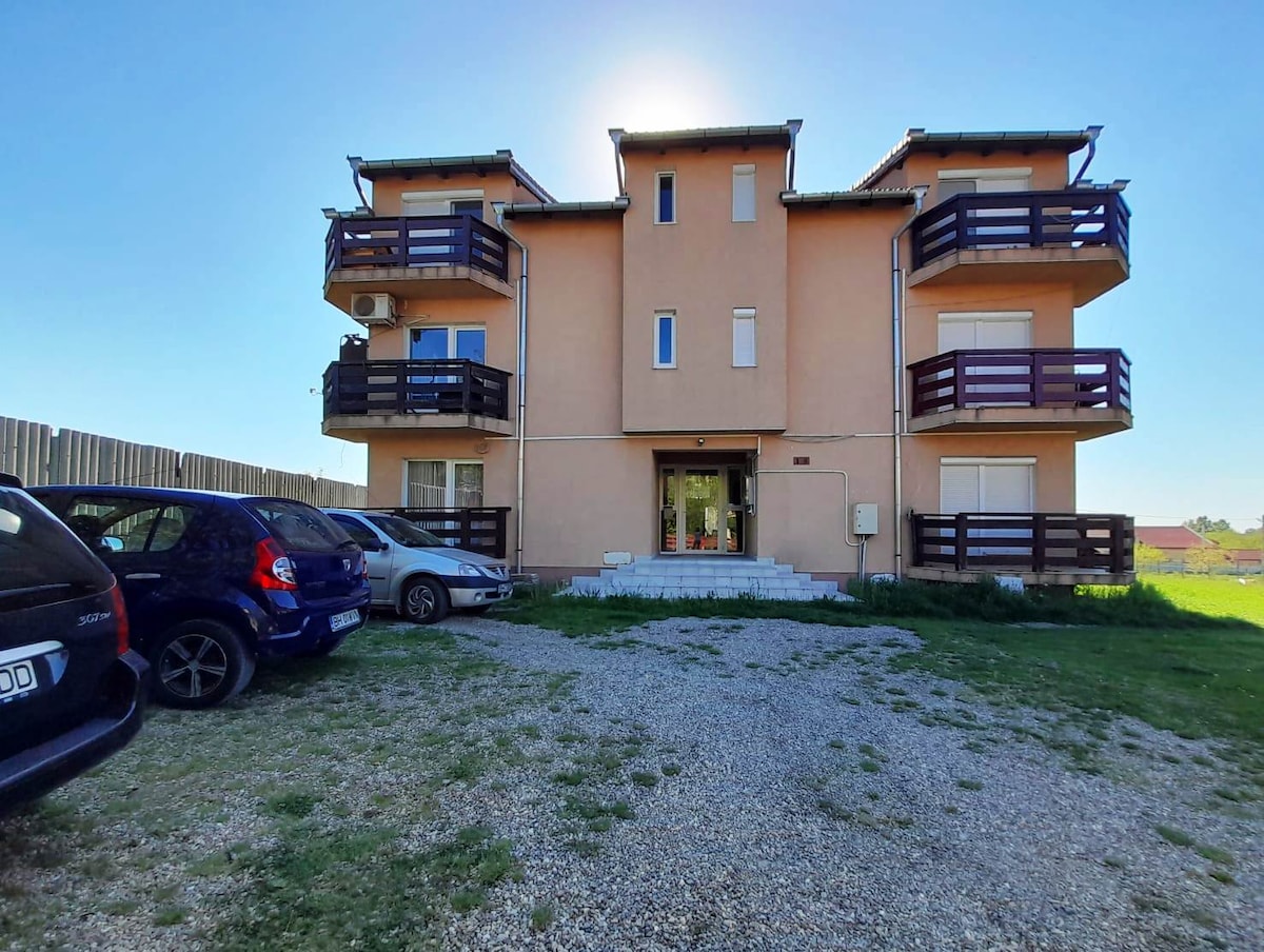 A modern building with a light exterior is shown, featuring three levels and multiple balconies. An open area with gravel and grass is visible in front, where several parked cars are arranged. The entrance is highlighted by a clear pathway leading inside.