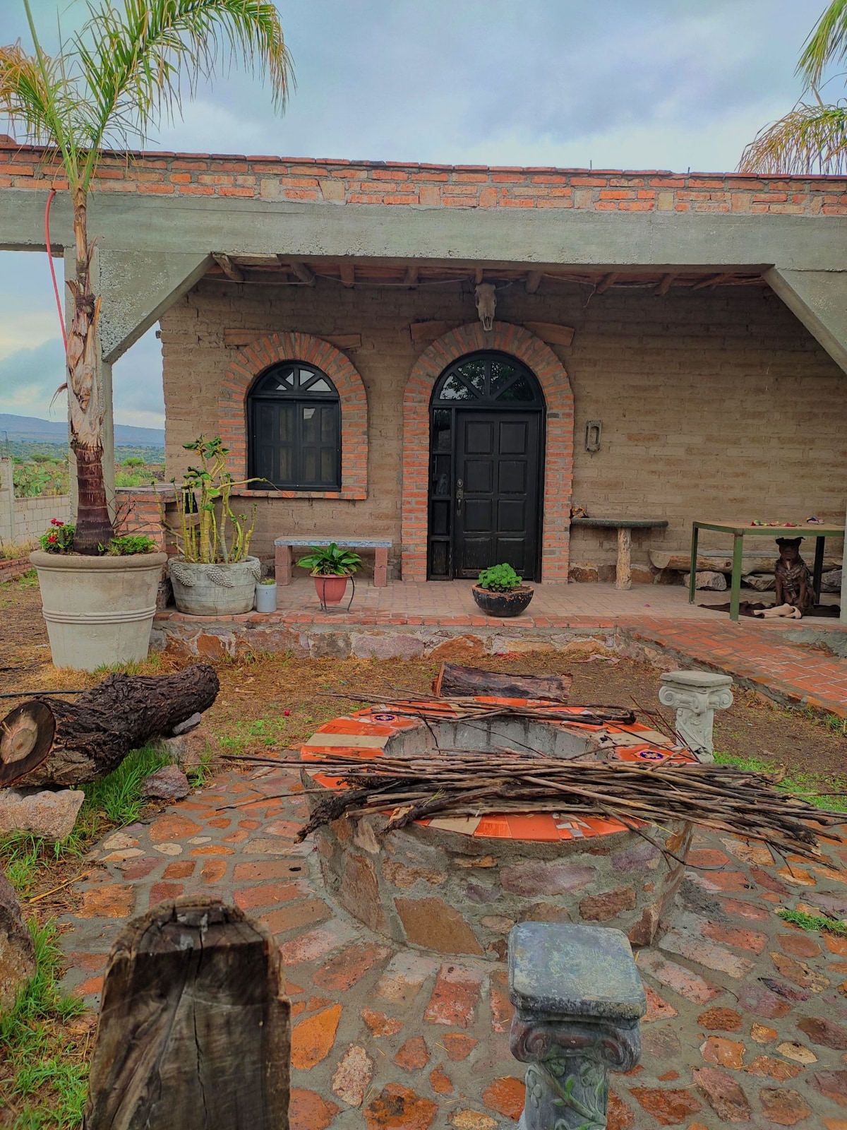 The image depicts a charming exterior with a stone and brick design. A circular fire pit is surrounded by stone seating, while potted plants are placed on the patio. Large arched windows and a solid black door enhance the rustic aesthetic of the structure.