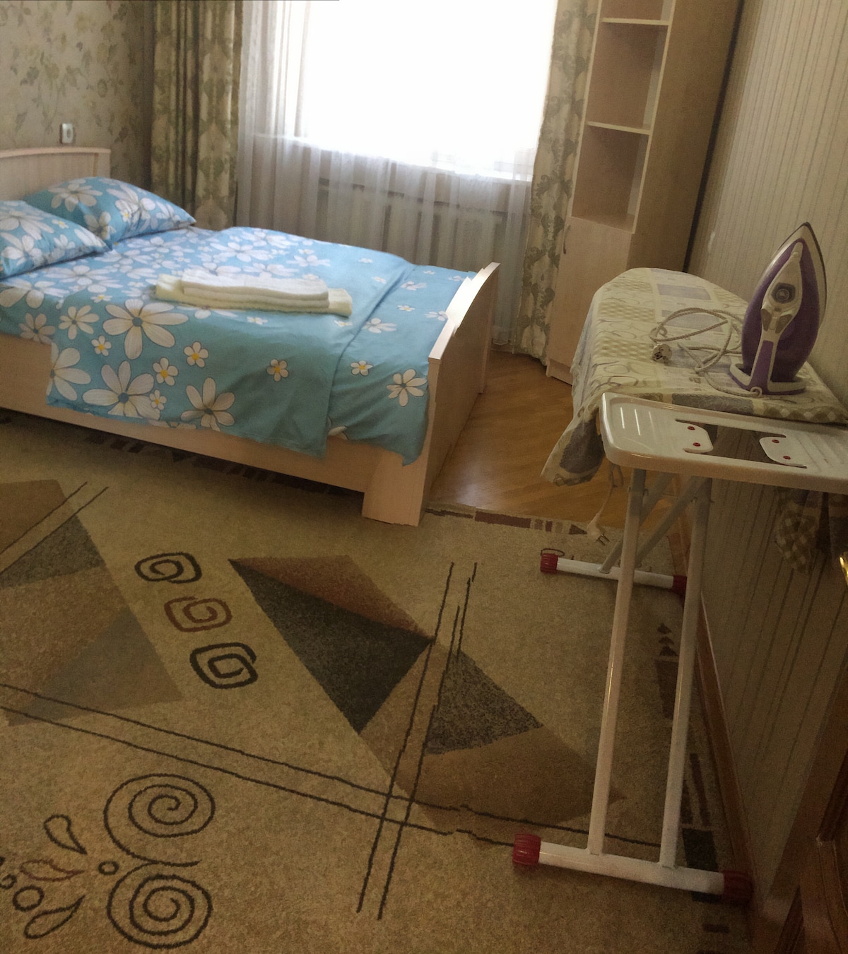A well-lit bedroom is featured, showcasing a spacious bed adorned with a floral-patterned blue bedspread. A neatly arranged set of towels is placed on the bed. An ironing board with an iron is positioned nearby, with a patterned area rug covering the wooden floor.