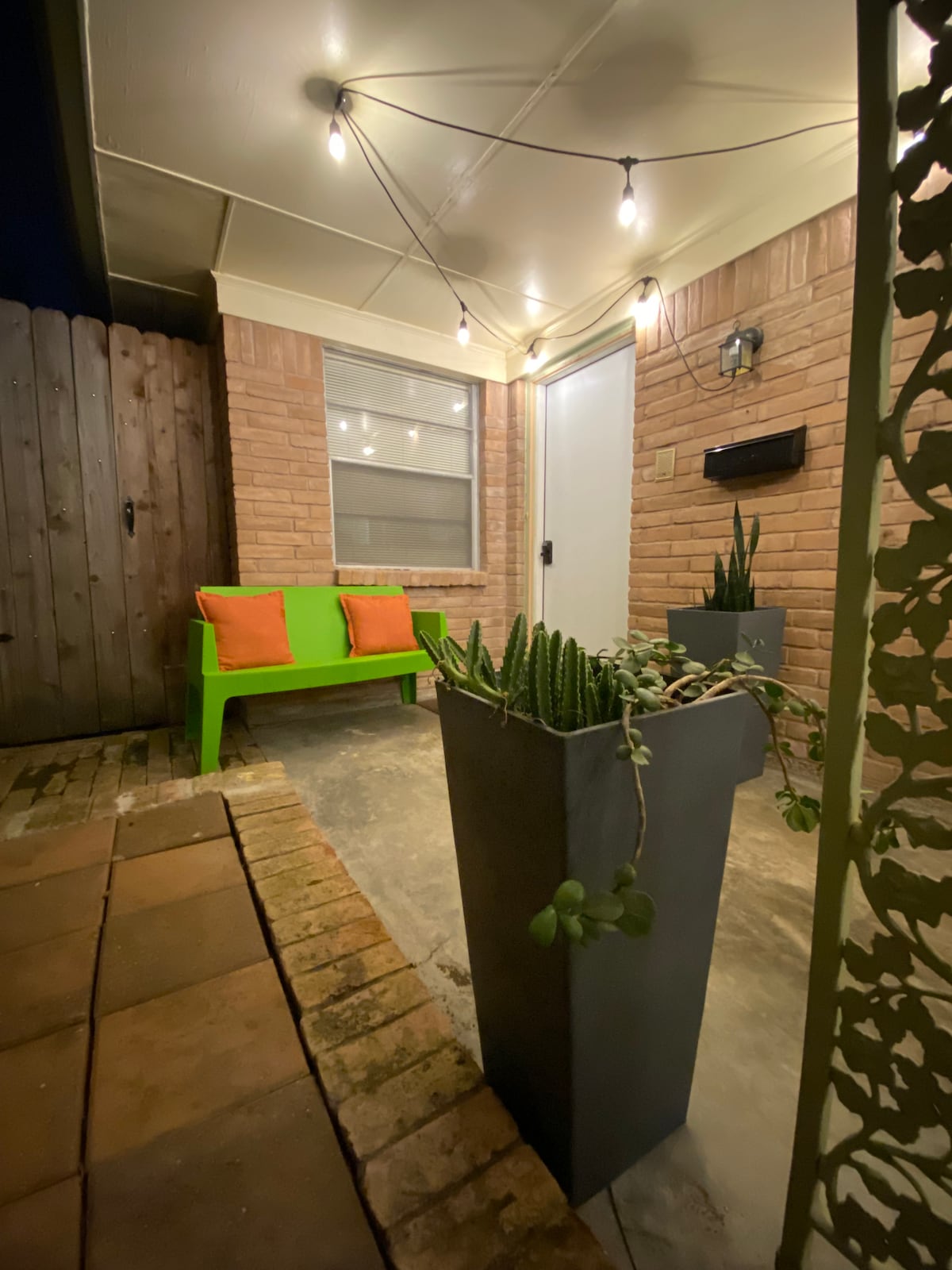 A welcoming entrance features a green seating area with two orange cushions. Planters with various plants are positioned beside the entrance, and soft lighting from string lights enhances the ambiance. The front door, framed by brick walls, leads into the duplex.