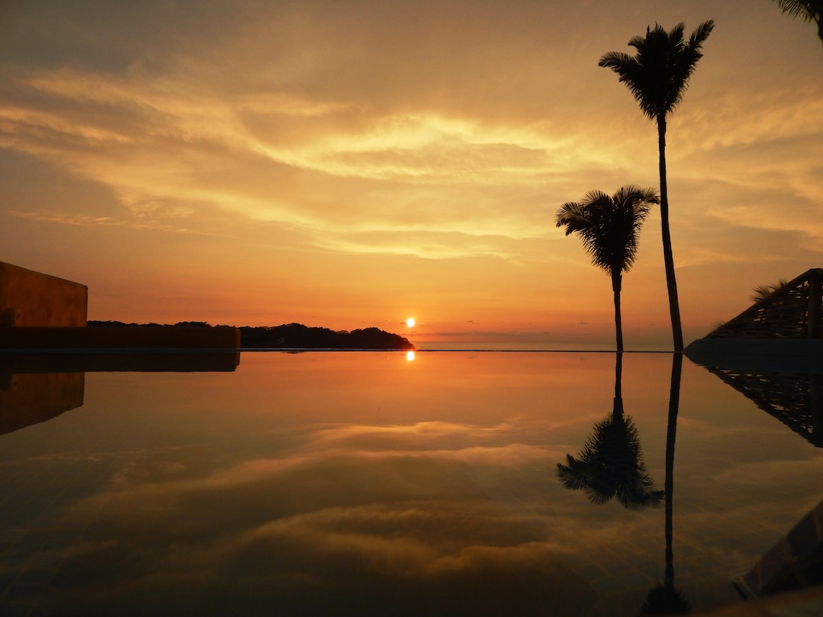 A tranquil sunset is reflected in the calm waters of the rooftop pool, surrounded by palm trees. The warm hues of orange and pink fill the sky, creating a serene atmosphere that invites relaxation.