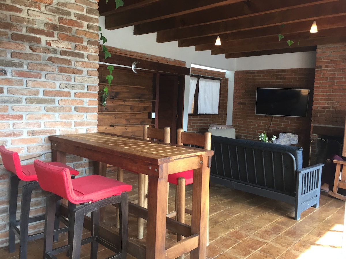 A spacious common area features a wooden dining table with red stools, complimented by a comfortable dark sofa. Exposed brick walls and wooden beams create a rustic ambiance, while a television is positioned for entertainment. Natural light enters through the nearby window, enhancing the inviting environment.