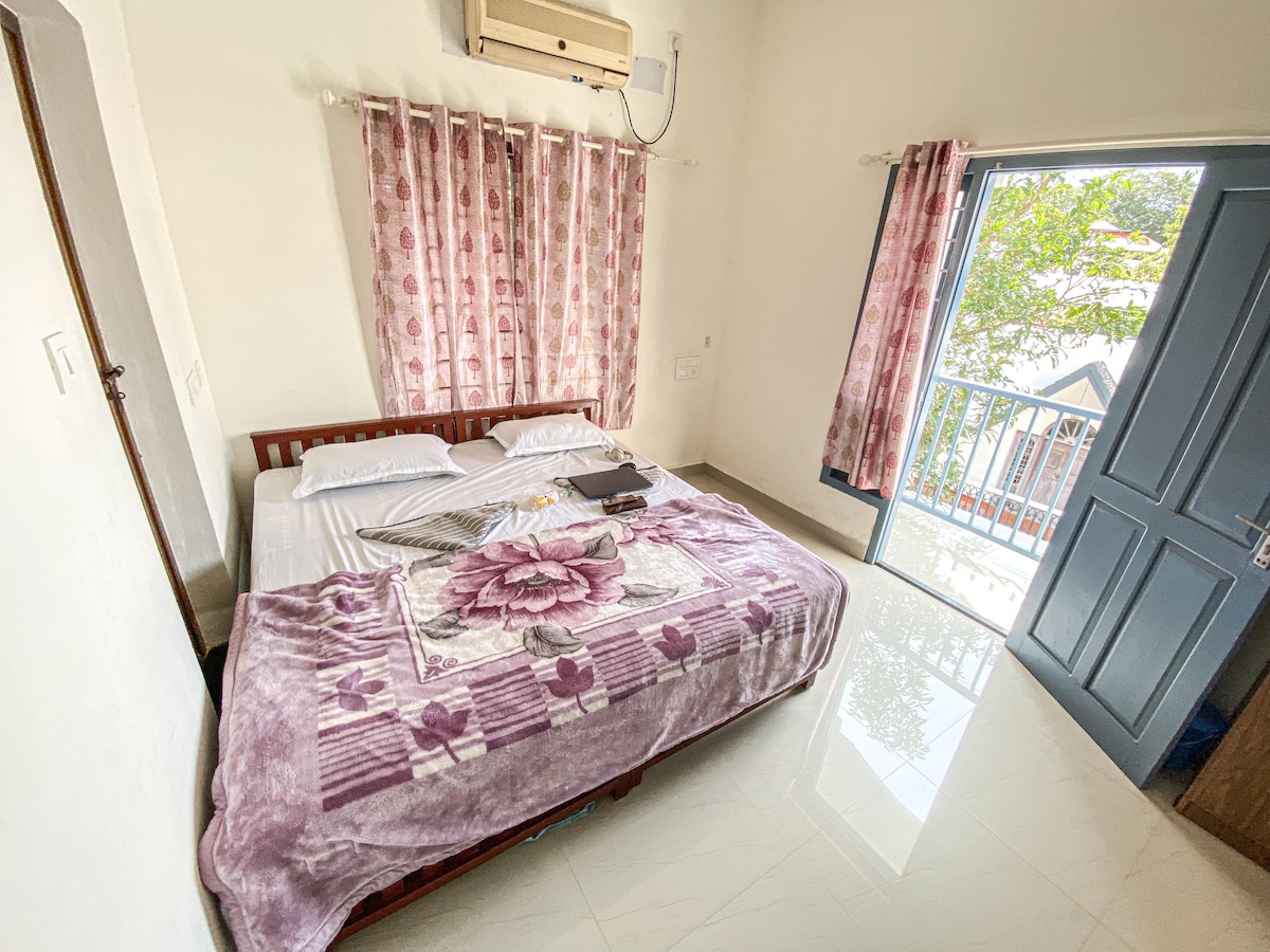 A bright room is furnished with a spacious bed covered by a floral blanket. Large windows with sheer curtains allow natural light to fill the space. An air conditioning unit is mounted on the wall, and a door leads to a small balcony, offering a view of the surroundings.