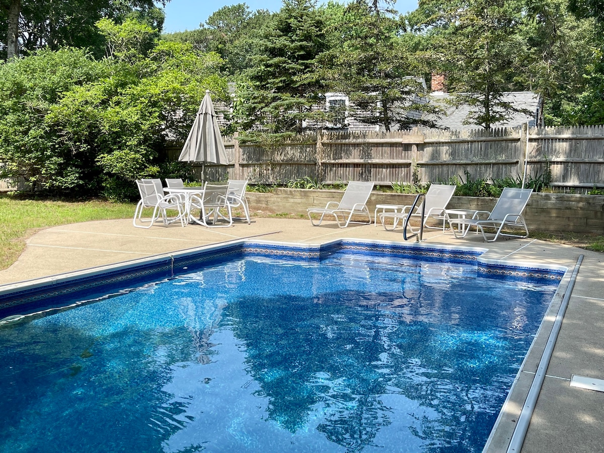 A built-in swimming pool is surrounded by a spacious patio area, featuring several lounge chairs and a dining table with an umbrella. Lush greenery and a wooden fence create a private outdoor space, inviting relaxation by the water.