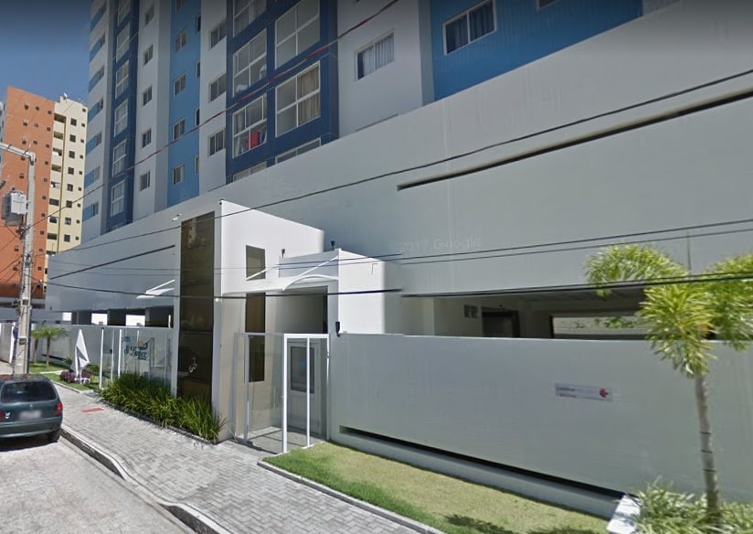 The exterior of a modern apartment building is showcased, featuring a sleek facade with glass elements. Low-maintenance landscaping lines the entrance, and parked cars are visible along the street. The structure rises several stories high, suggesting a well-maintained community atmosphere.