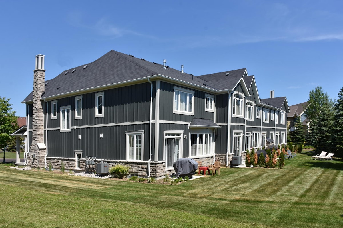 The exterior of the townhome showcases a modern design with dark wood siding and stone accents. A well-manicured lawn with neatly trimmed grass surrounds the home. The patio area features outdoor furniture and a propane grill, providing a comfortable space for relaxation.