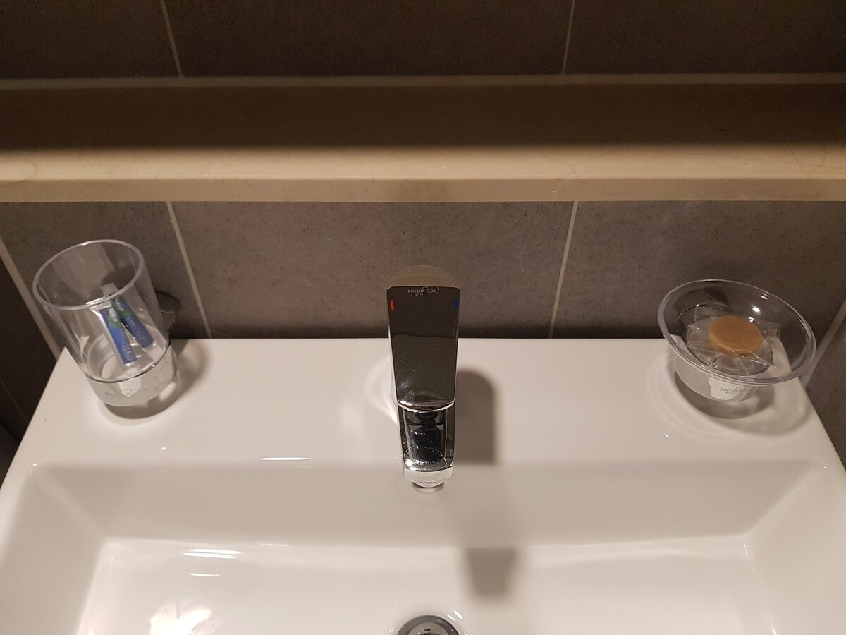A clean and modern bathroom sink is showcased, featuring a sleek faucet positioned at the center. Two transparent containers on either side hold toiletries, while a smooth, white countertop provides ample space. Neutral-toned tiles complement the minimalist design.