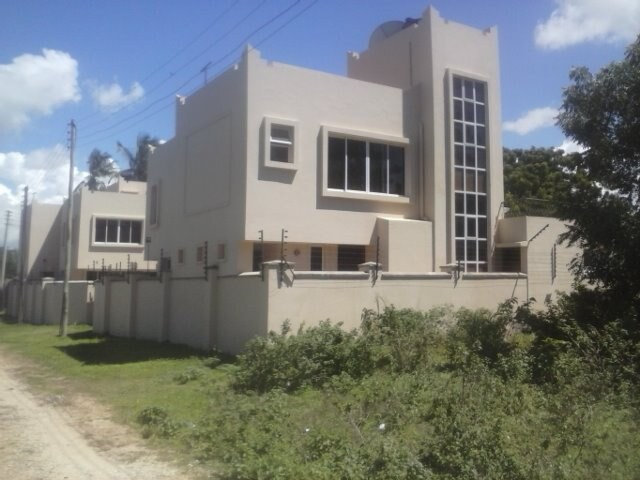 A modern two-story building is shown, featuring large windows that provide natural light. The exterior is painted in neutral tones, and a concrete perimeter wall is visible. Surrounding greenery adds to the serene setting, along with utility poles lining the road.