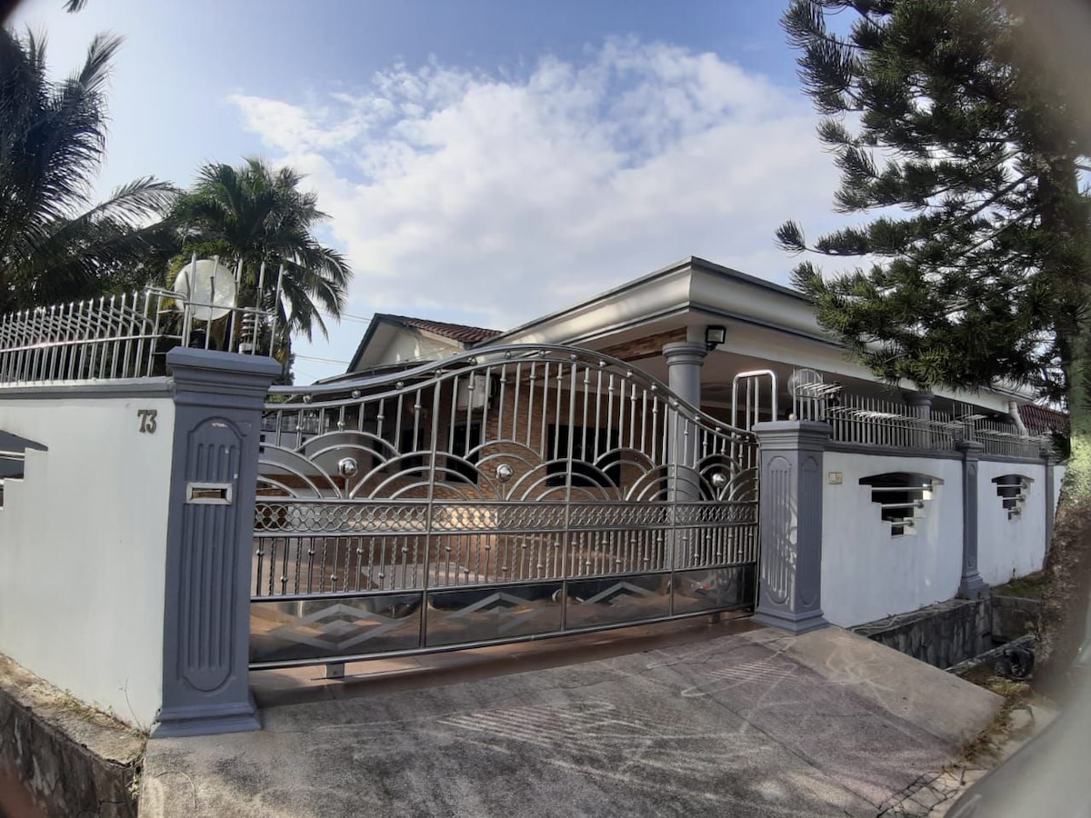 A modern entrance gate is visible, featuring a decorative design and flanked by palm trees. The path leads to a brick house with a low-pitched roof, under a partly cloudy sky. Surrounding greenery enhances the welcoming appearance.