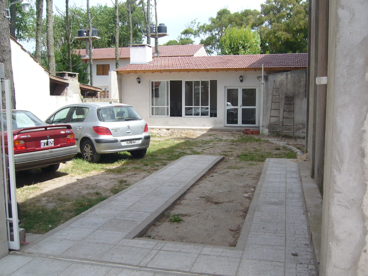 A spacious outdoor area is visible, featuring a gravel and tiled pathway leading to a modern structure with large glass doors. Two parked vehicles, a red sedan and a silver hatchback, are located nearby amidst a grassy setting. Surrounding greenery provides a serene backdrop.