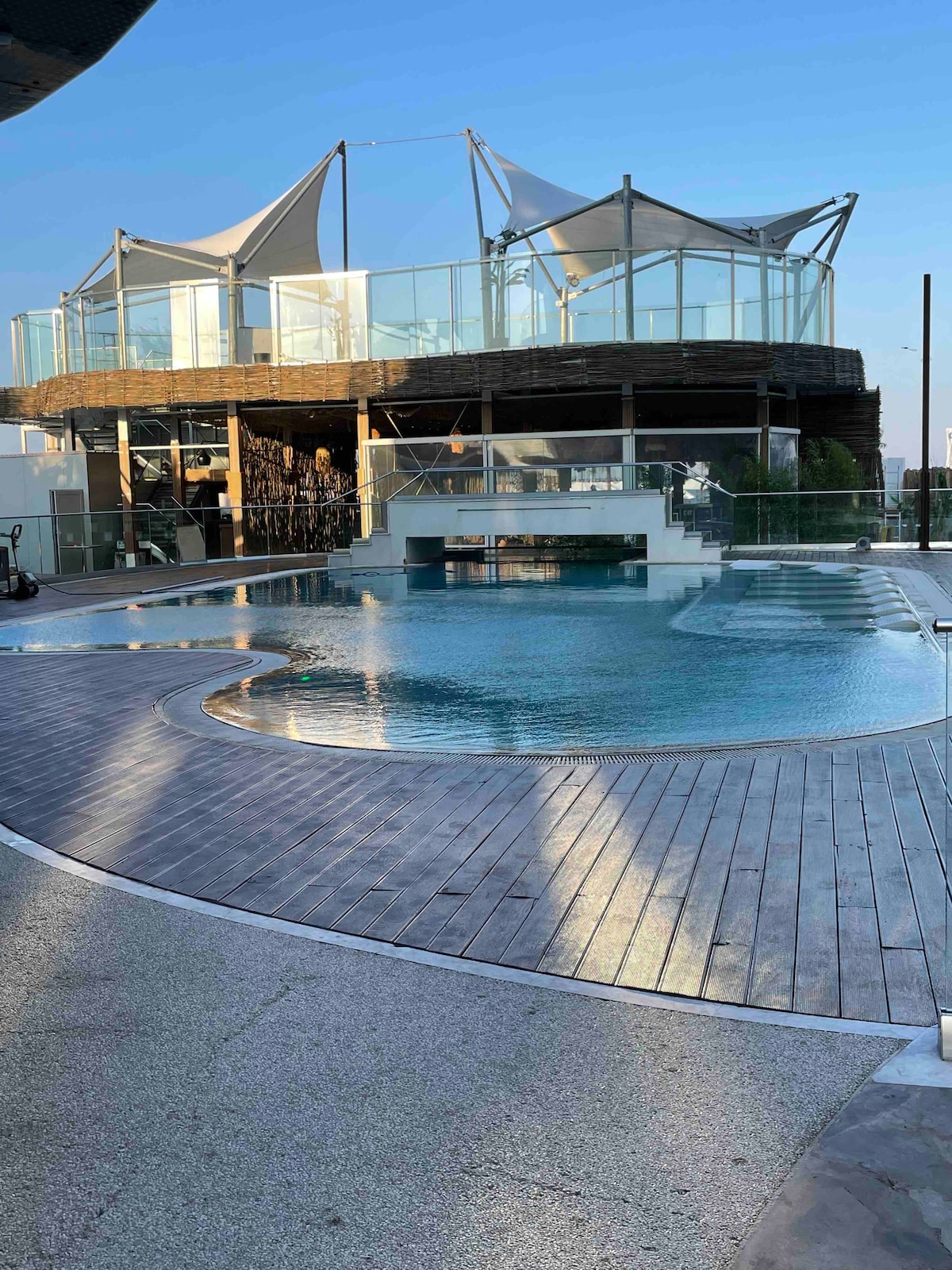 A rooftop pool area is presented, featuring a curved design and surrounded by wooden decking. The clear water reflects the sky, while a modern structure with shaded seating areas overlooks the pool. Lush greenery is visible in the background, enhancing the overall ambiance.