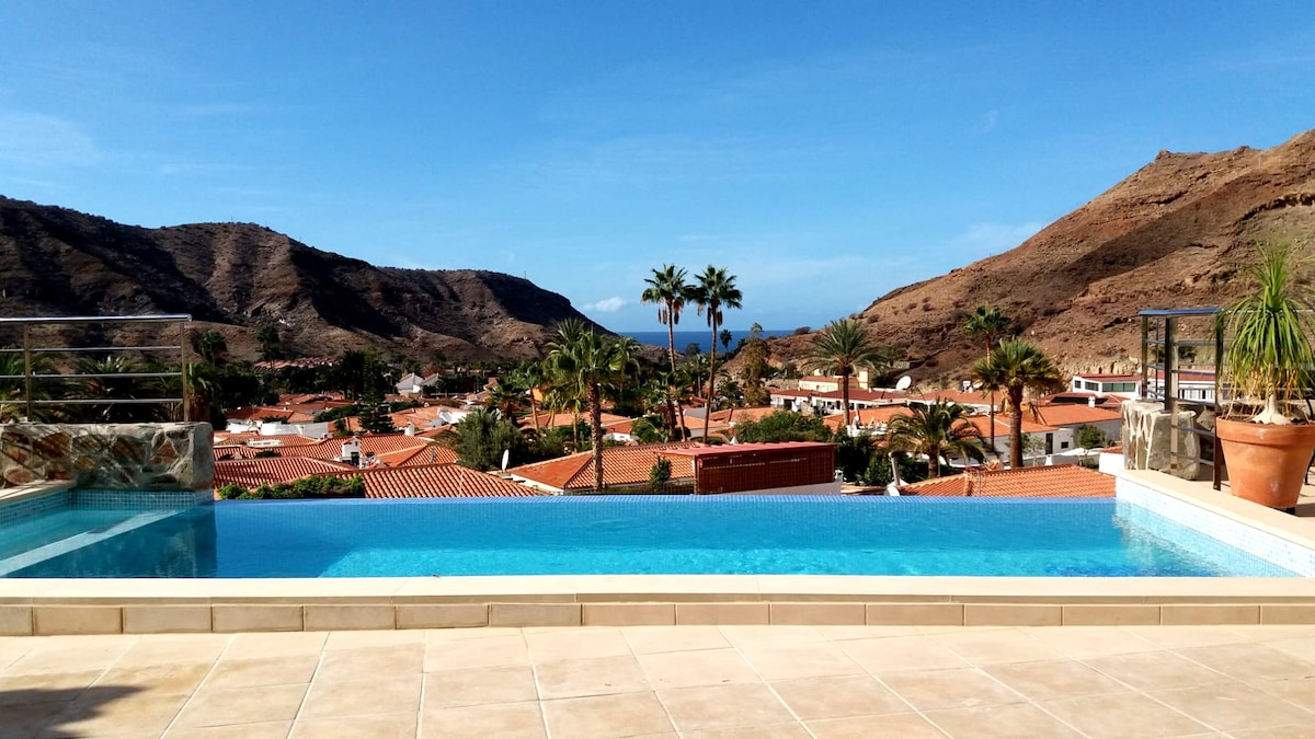 An infinity pool extends into the horizon, offering uninterrupted views of the Tauro valley and the Atlantic Ocean. Surrounding landscape includes terracotta rooftops and palm trees, complemented by a backdrop of rugged mountains under a clear blue sky.