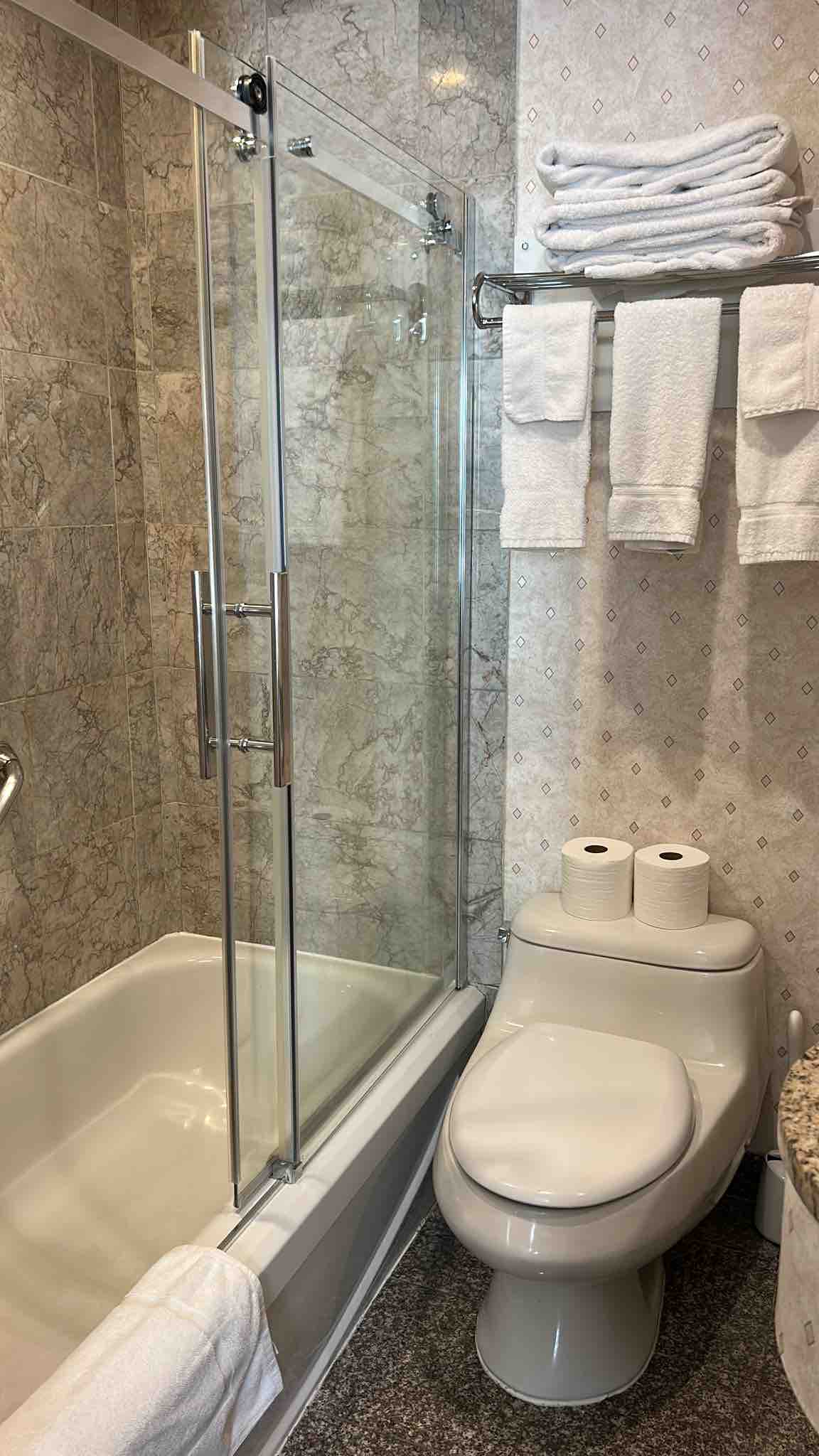 The bathroom features a glass-enclosed shower adjacent to a bathtub, complemented by marble walls. Clean towels are neatly arranged on a rack, and two rolls of toilet paper are visible on the countertop. A modern toilet completes the well-appointed space.