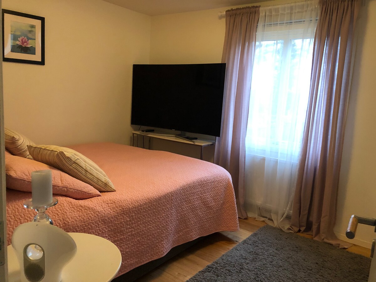 A spacious bedroom features a large television positioned opposite a cozy bed adorned with a soft pink coverlet. Natural light filters through a window draped with sheer curtains, while a small side table holds a candle, adding to the room's tranquil ambiance.
