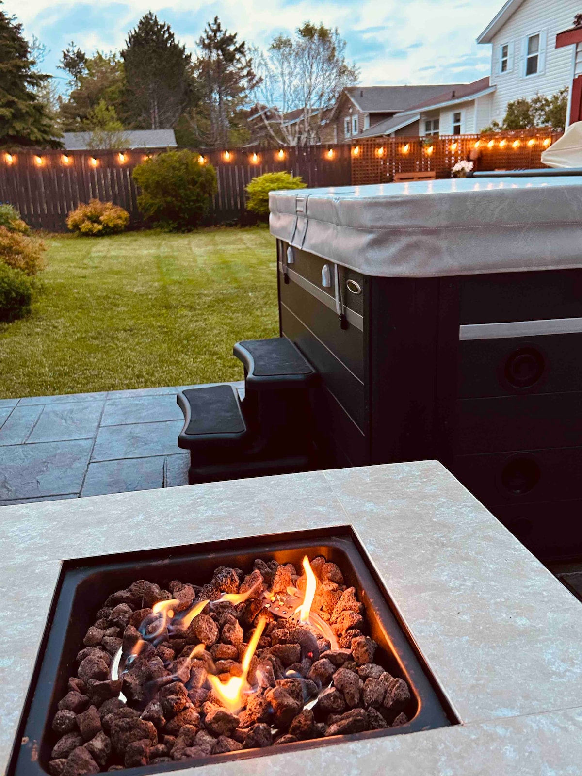A cozy outdoor space is highlighted by a propane fire table, with flickering flames visible among decorative rocks. In the background, a newly installed hot tub sits near a well-maintained lawn, surrounded by string lights that illuminate the area, creating a welcoming ambiance.