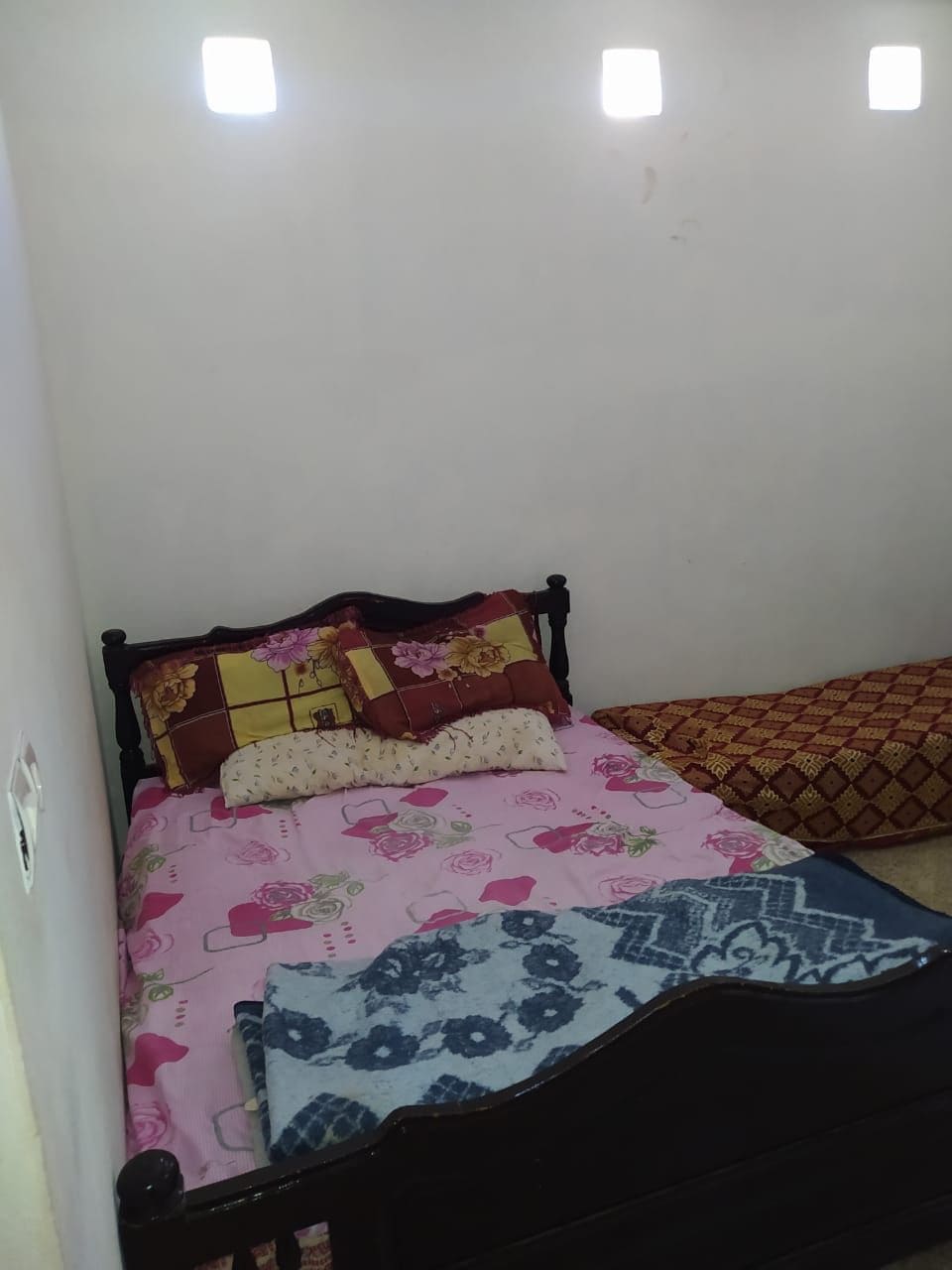 A simple bedroom features a sturdy black bed adorned with floral-patterned bedding. Decorative pillows are positioned against the headboard. A patterned blanket is draped across the bed, while a cushioned mat sits nearby. Natural light filters in through small wall windows.