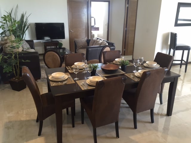 A dining area features a dark wooden table set for eight, complemented by brown upholstered chairs. A small decorative plant sits at the center. In the background, a living area is visible with a sofa, coffee table, and a television against a wall.