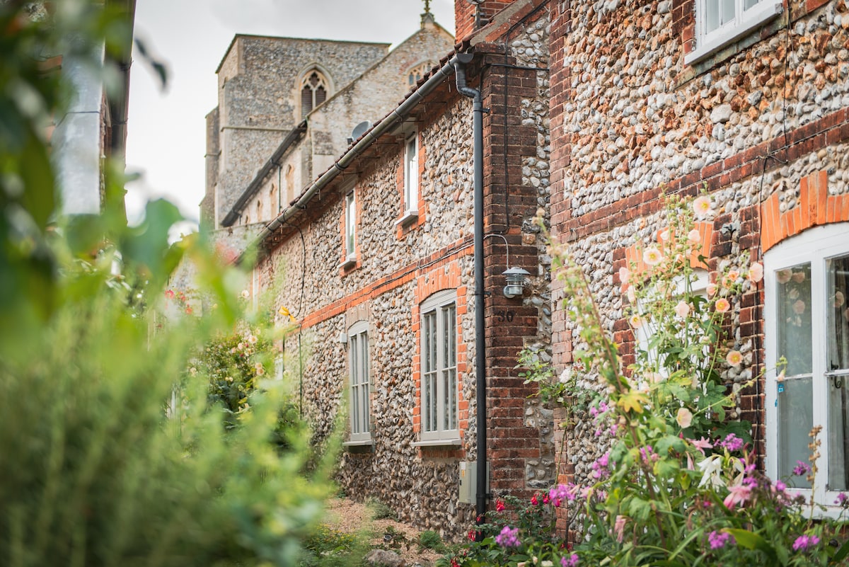 A traditional flint cottage is surrounded by vibrant blooming flowers, with its rustic exterior featuring a blend of flint and brick. The windows are framed with white trim, reflecting a charming countryside aesthetic. A glimpse of a medieval church can be seen in the background.