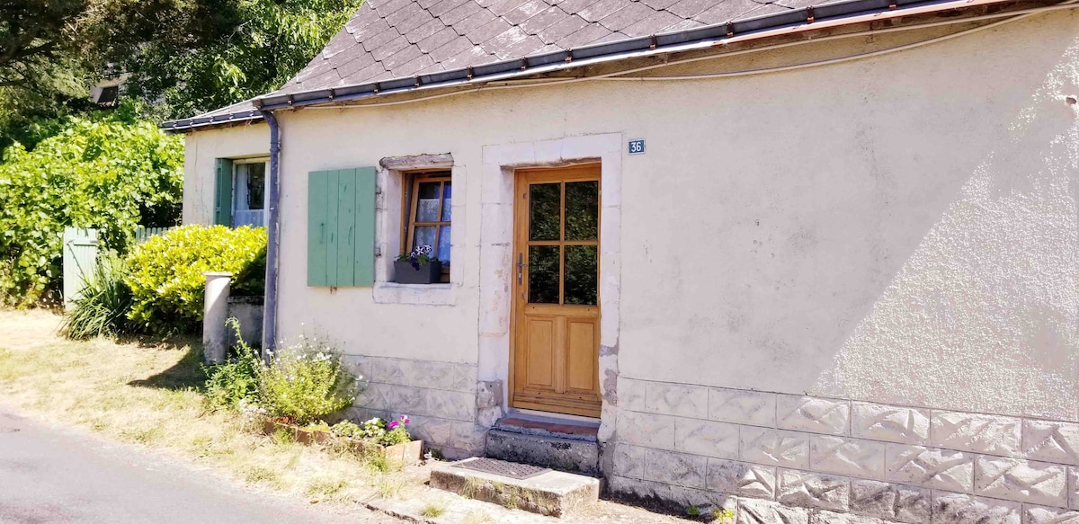 The cottage features a charming façade with light-colored stone walls and green shutters. A wooden door with glass panes stands at the entrance, framed by a small step. Lush greenery surrounds the home, enhancing the inviting exterior.