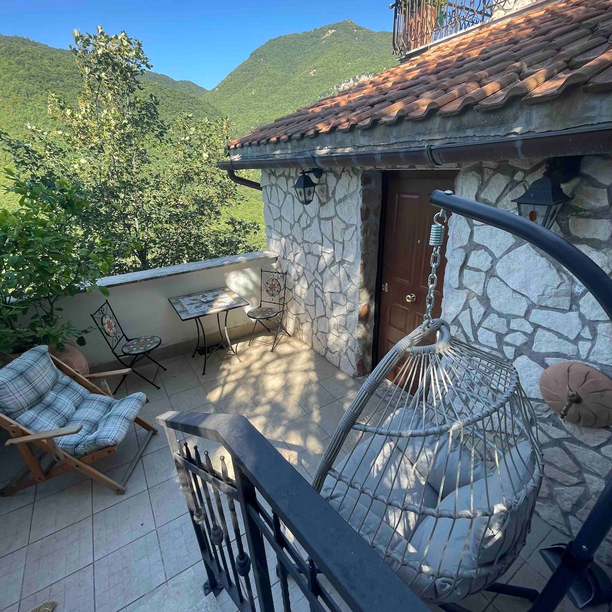 A charming terrace is featured, with a hanging chair and two seated patio chairs beside a small table. The stone wall structure is complemented by views of the surrounding green mountains, with sunlight creating gentle shadows across the tiled floor.