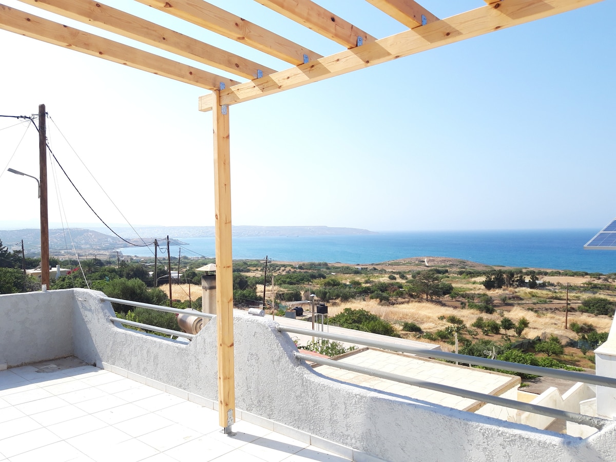 A spacious terrace with wooden beams offers an unobstructed view of the sea and surrounding landscape. The low, white wall creates a boundary while allowing for a panoramic vista of the coastline and distant hills under a clear blue sky.