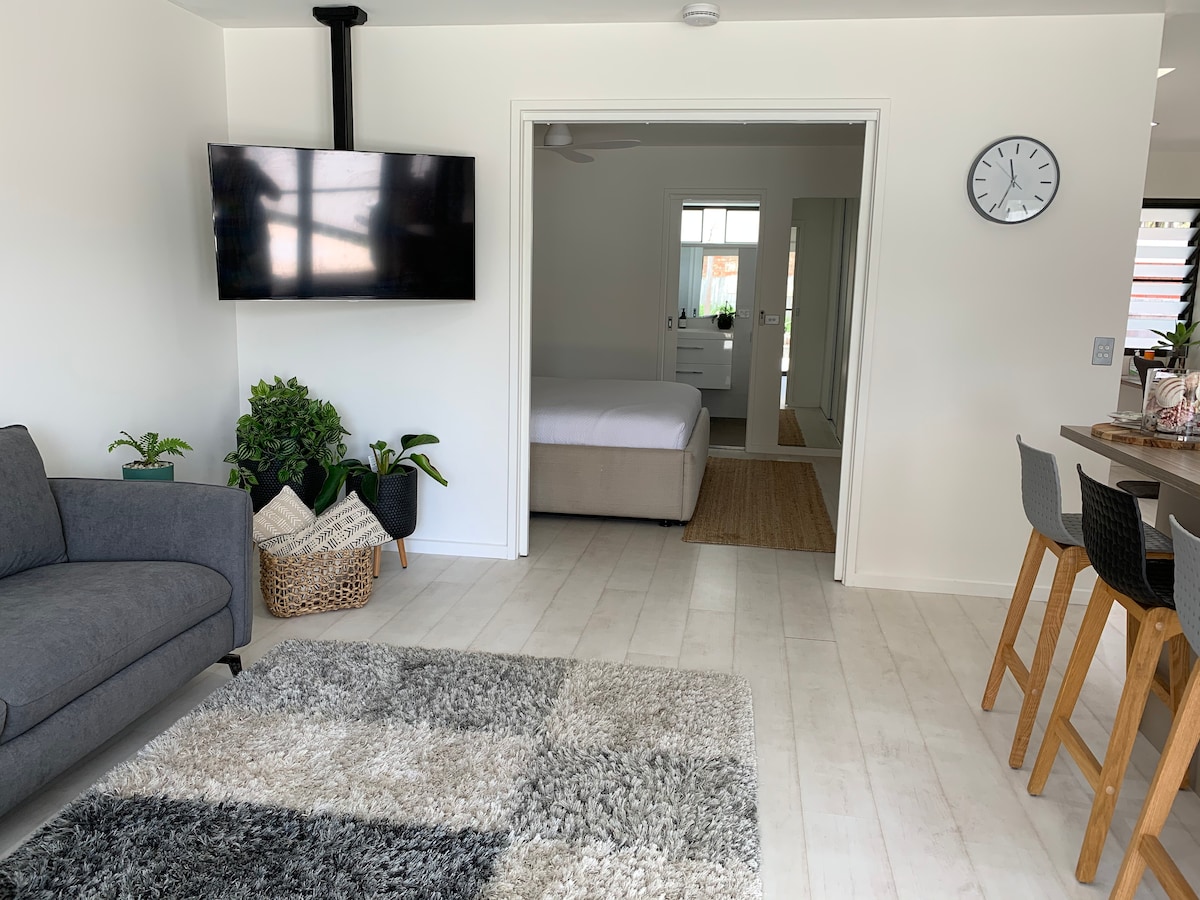 A spacious living area is furnished with a gray couch and a dining table with tall chairs. A wall-mounted television is positioned for viewing. Light streams through multiple windows, while a textured rug adds warmth to the floor. A cozy bedroom is visible through the open doorway.