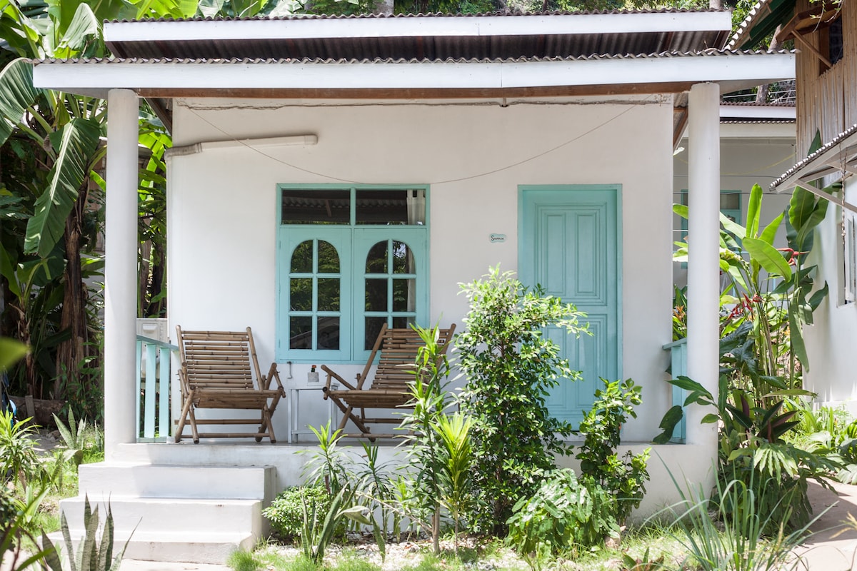 A bungalow exterior featuring light blue accents, with two wooden lounge chairs placed on a small porch. Lush greenery surrounds the structure, providing a tropical ambiance. A door and large windows allow for natural light to enter, enhancing the inviting character of the space.