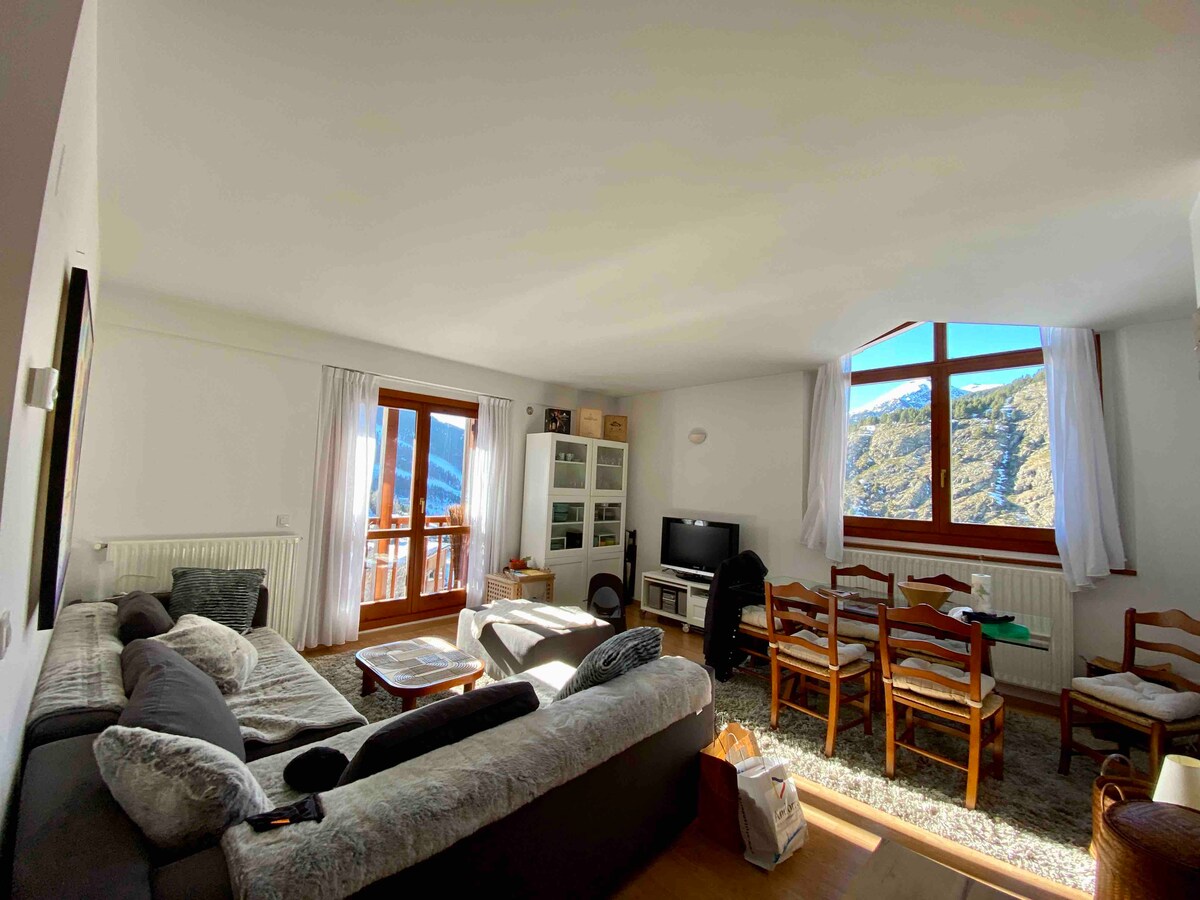 A spacious living area features a large sectional sofa and a light rug, complemented by a wooden dining table with chairs. A large window overlooks the scenic mountains, allowing ample natural light to enter. A TV is positioned near a cozy storage unit.