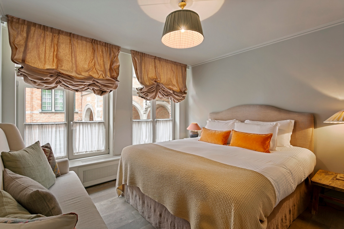 The bedroom features a spacious bed with luxurious bedding, accented by warm orange pillows. Large windows are adorned with soft, flowing curtains, allowing natural light to brighten the room. A comfortable seating area is positioned nearby, enhancing the room's inviting atmosphere.