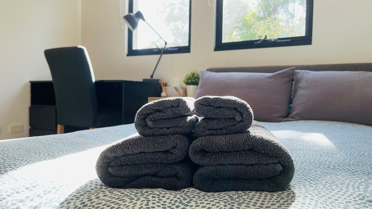 A neatly arranged stack of dark towels is positioned on a bed with a textured coverlet. In the background, a small desk with a chair and a lamp can be seen near a window, allowing natural light to illuminate the space.