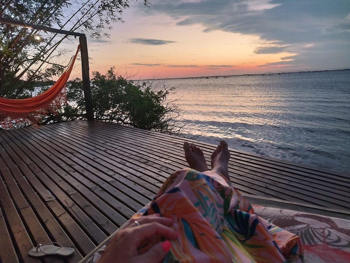 A scenic view of a tranquil shoreline is captured at sunset, with soft colors reflecting on the water. A wooden deck is shown, adorned with a hammock and tropical foliage nearby, providing a serene setting for relaxation.