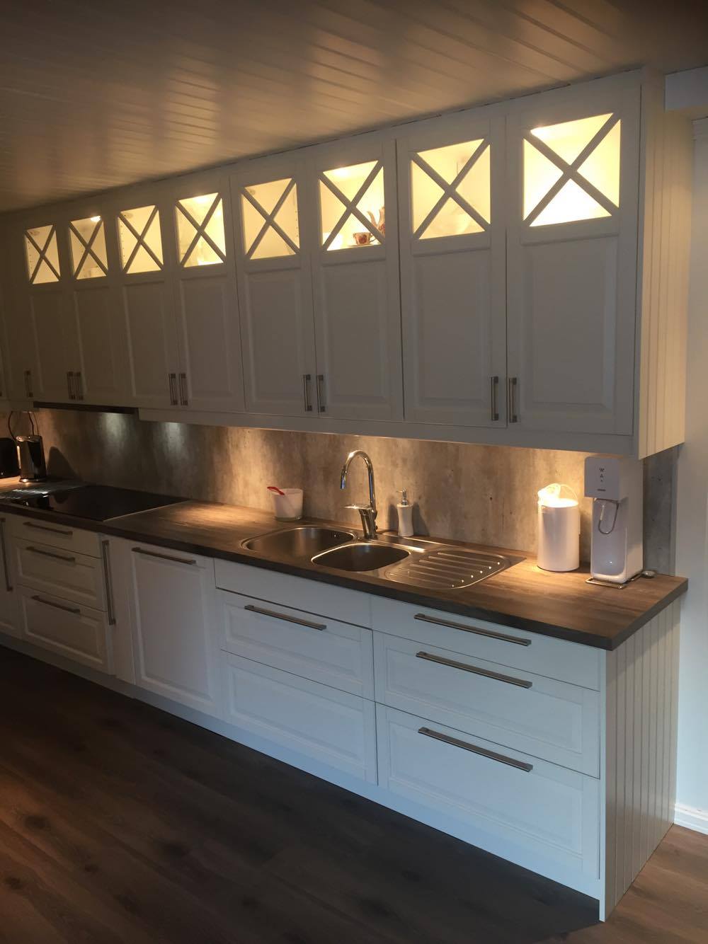 The kitchen features white cabinetry with glass-paneled doors, complemented by a neutral countertop. A double sink is positioned beneath cabinet lighting, illuminating the workspace. Appliances such as a kettle and coffee maker are visible, set against a backdrop of textured wall panels.