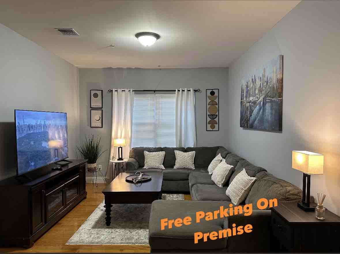 A spacious living area is equipped with a large sectional sofa and a wooden coffee table. A flat-screen television is mounted on the wall, accompanied by two side lamps providing gentle illumination. Curtains frame the window, while artwork enhances the room's decor.