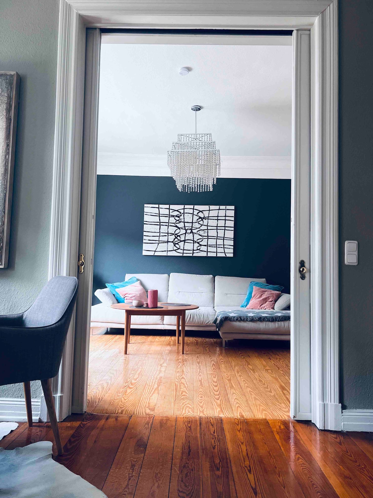 A spacious living area is visible through an open doorway. A light-colored sofa is accompanied by a coffee table, with decorative pillows adding color. A contemporary art piece adorns the dark blue wall, while a chandelier provides overhead lighting.