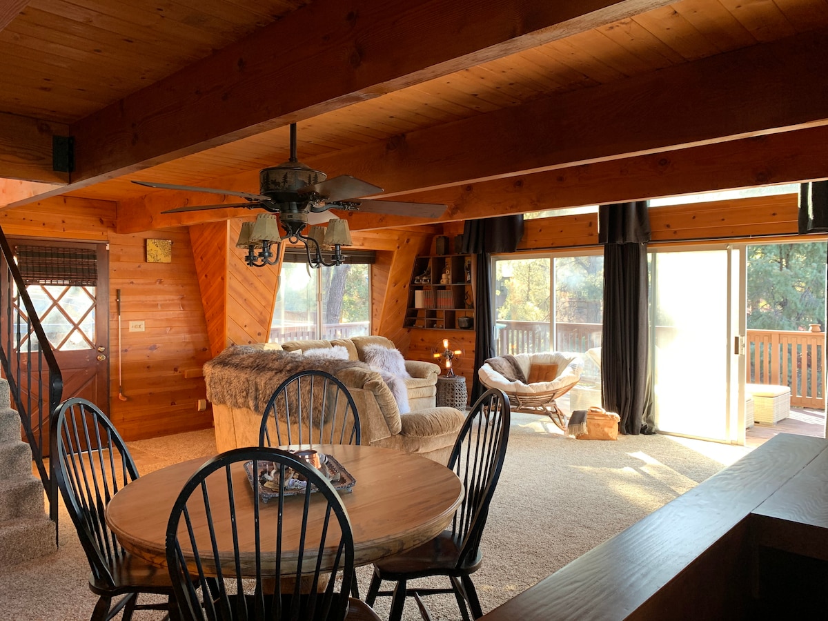 An open floor plan showcases a cozy living area with large windows allowing natural light to fill the space. A wooden dining table is paired with black chairs, while a comfortable sectional couch and papasan chair provide inviting seating options. A sliding door leads to the outdoor deck.