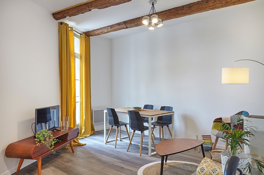 A bright living area features wooden beams overhead, with large windows draped in yellow curtains. A rectangular dining table is surrounded by dark chairs, while a modern television is mounted on a sleek wooden console. A cozy seating area is accentuated by a floor lamp and decorative plant.