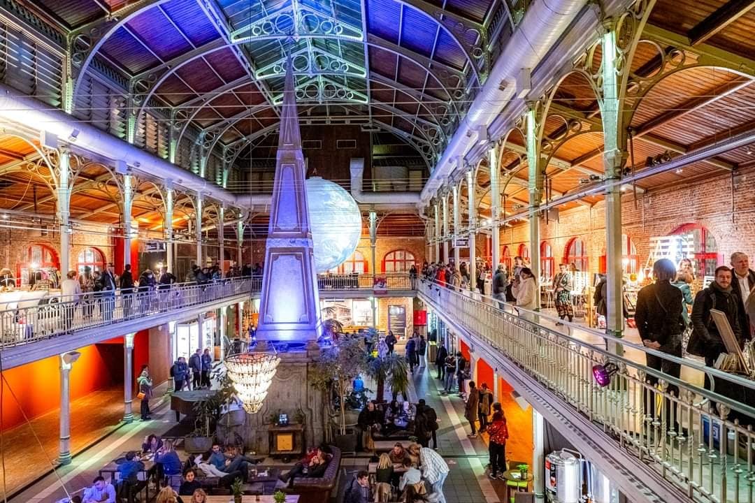 An expansive indoor market space is depicted, featuring a high arched ceiling and decorative ironwork. Various vendors occupy the stalls, with guests mingling throughout the area. A large chandelier hangs above a central seating area lit by soft lighting, creating a lively yet inviting environment.