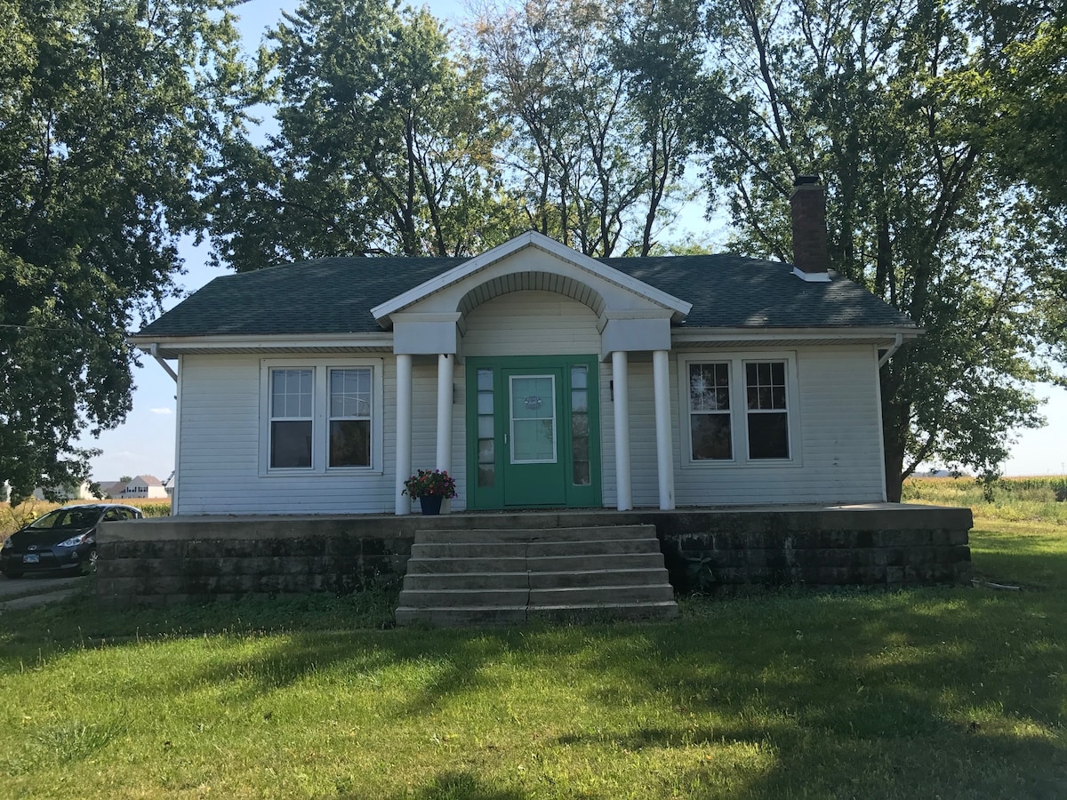 A charming farmhouse exterior is presented, featuring a green front door framed by white columns. Large windows allow natural light, and a stone staircase leads to the entrance. Surrounding greenery adds a touch of nature to the inviting facade.