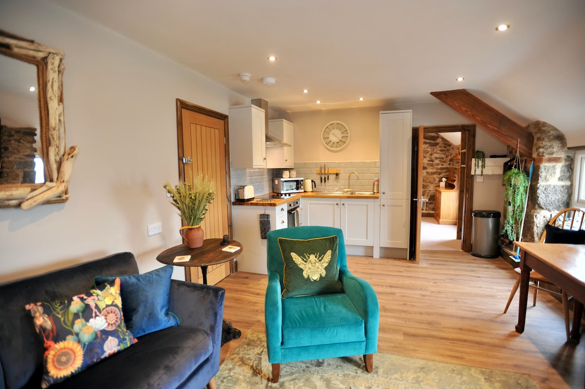 The open-plan living area displays a blend of modern and rustic design, featuring a cozy blue chair and a small round table. The fully fitted kitchen includes white cabinetry and stainless steel appliances. Exposed stone walls add texture to the space, complemented by warm wood accents.