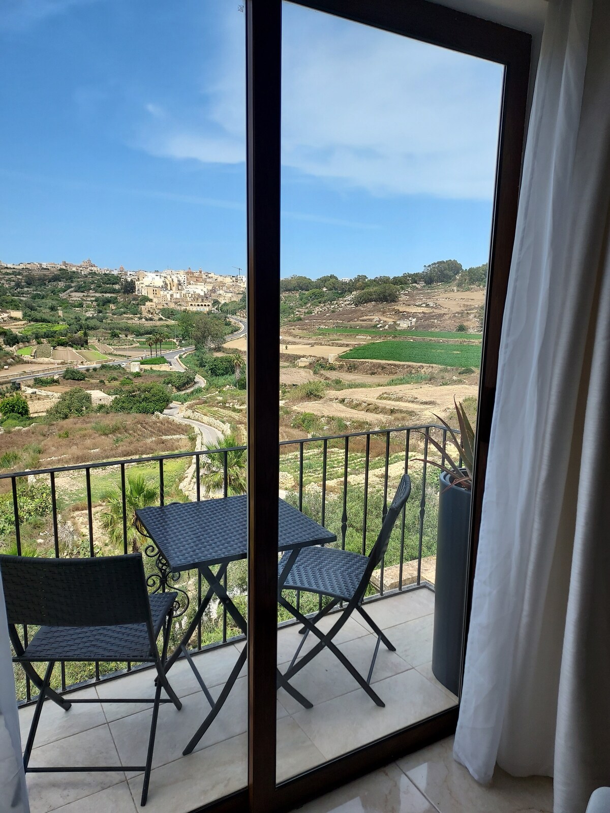 A balcony view showcases a serene countryside landscape, featuring manicured fields and rolling hills under a clear blue sky. Two chairs and a small table are positioned on the balcony, inviting guests to enjoy the natural surroundings.