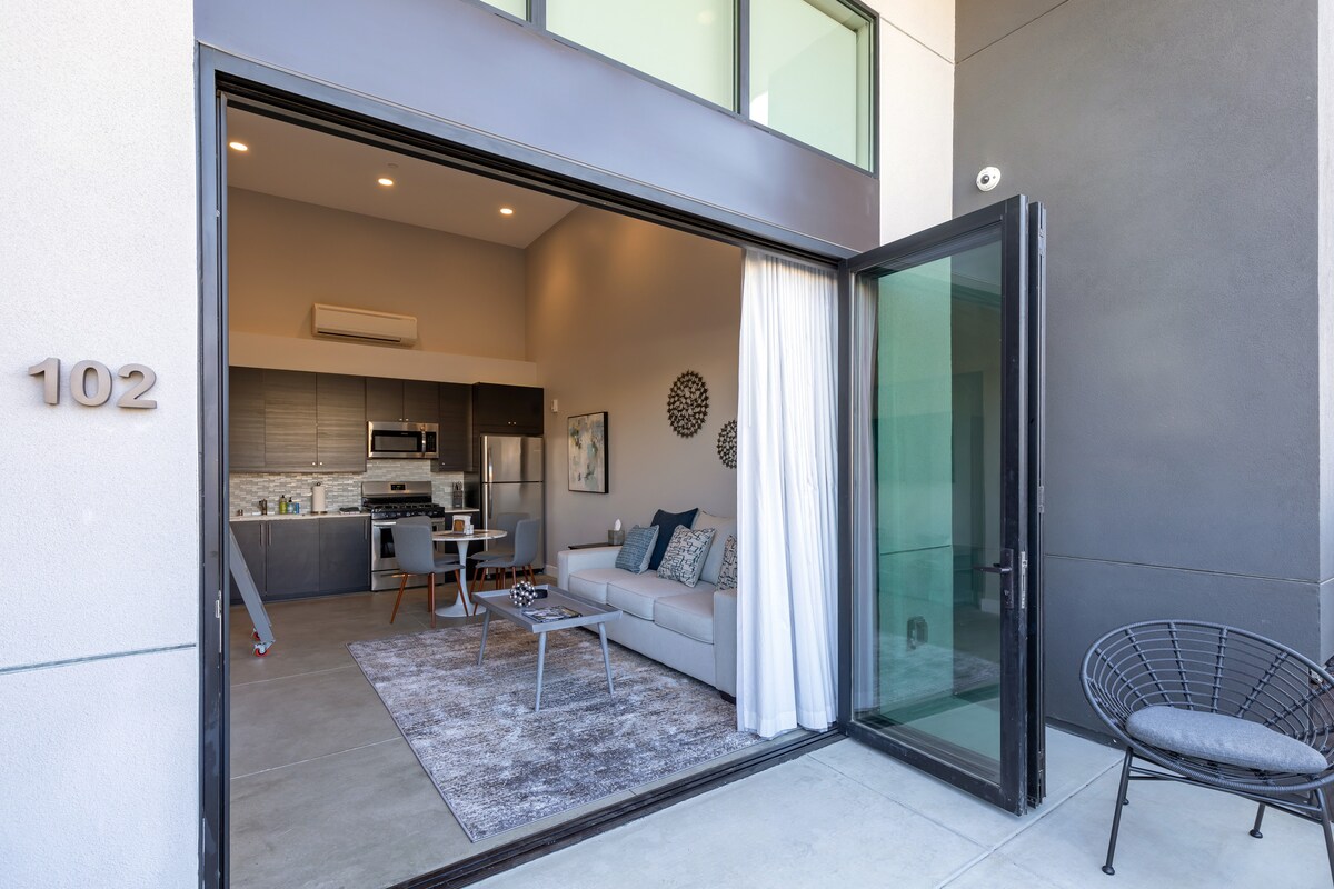 An open entryway reveals a modern living space with light-colored furniture. Accordion doors seamlessly connect the interior with the outside. A cozy rug is placed under a comfortable sofa, while a dining area is visible in the background, featuring sleek cabinetry and stainless steel appliances.
