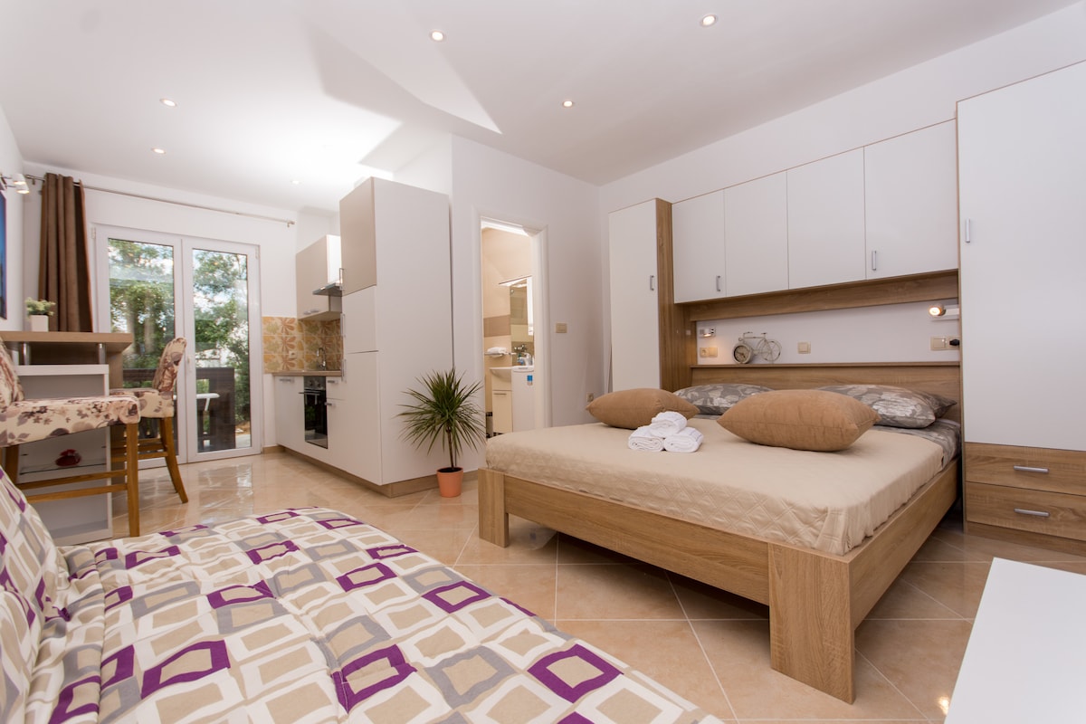 A spacious sleeping area features a large bed with soft pillows. Light-colored cabinetry complements the room, while a wooden table with a chair is positioned nearby. Natural light enters through sliding glass doors, enhancing the bright and airy ambiance.