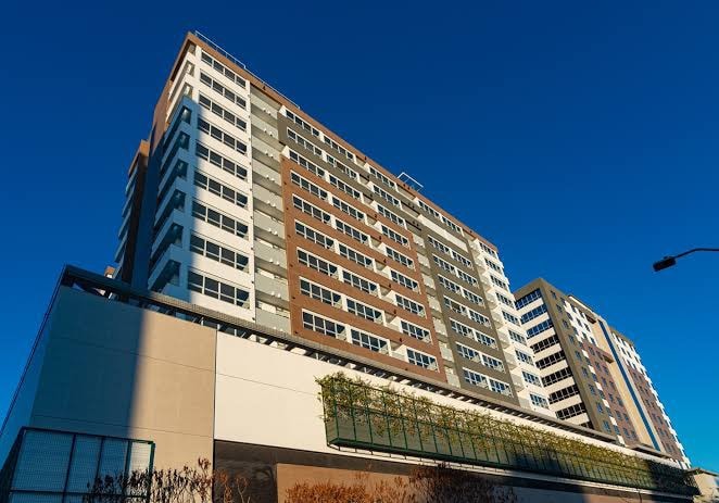 The exterior of a modern multi-story building is captured against a clear blue sky. The structure features a mix of light and dark facade elements, with large windows providing natural light. A green terrace runs along the base, enhancing the urban landscape.