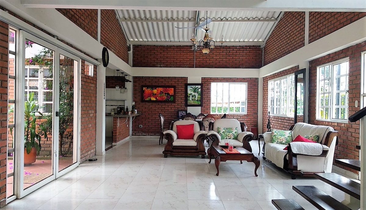 A spacious living area is highlighted by large windows that provide ample natural light. The room features comfortable seating arrangements, including a couch with decorative cushions. A dining space is visible, and the warm tones of the brick walls complement the bright decor.